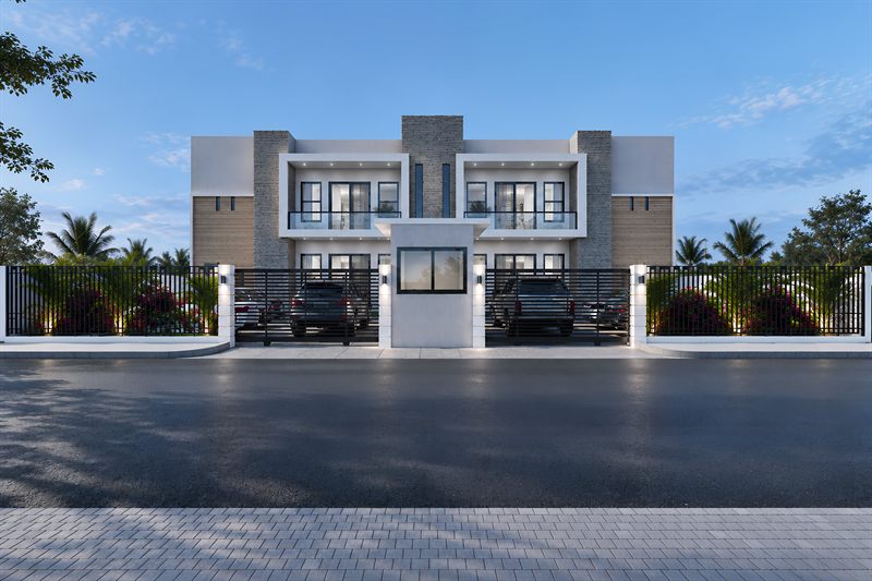 The block square on from the road: two storeys in white render with stone-clad piers and timber panels, balconies behind glass balustrades on the upper floor, a guard house in the middle of the front wall, and cars parked behind the railings between beds of bougainvillea.