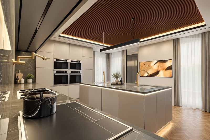 The kitchen from the hob end: the slatted timber ceiling running the length of the room, twin wall ovens set into a run of pale cabinetry, a brass pot-filler over the hob in the foreground, and the stone-topped island with a brass tap running out towards full-height glazing.