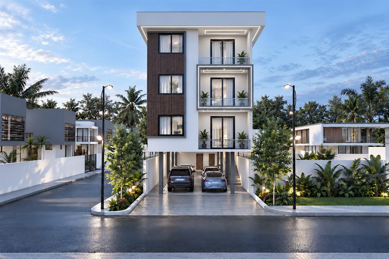 The block square on from the road: four floors in white render, three of flats with a timber-clad bay up the left of the front and stacked balconies at the centre, over a ground floor where two cars are parked between the columns, with street lamps and boundary walls either side.