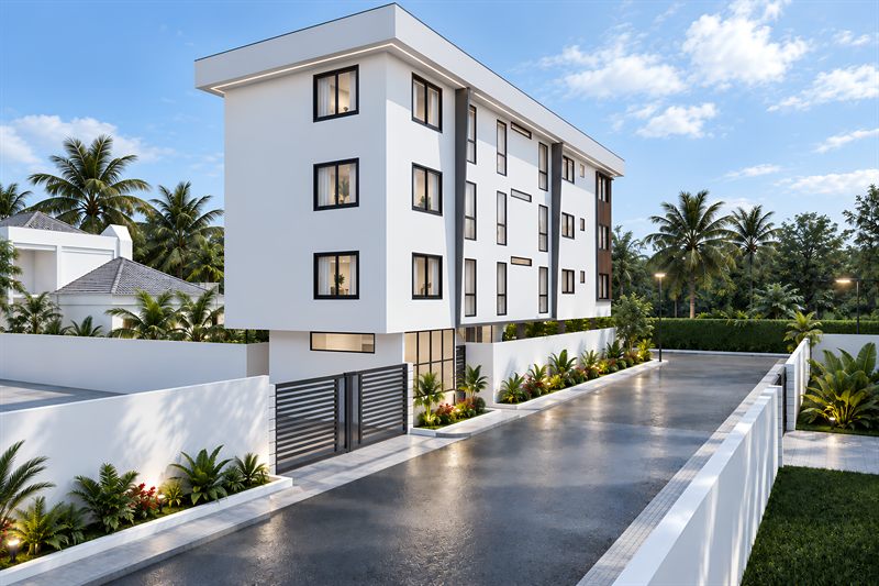 The block from the road at an angle, the flank running away with windows stacked over the three floors of flats, the security room glazed and lit at the back of the ground floor behind its planting bed, and a slatted gate set in the white boundary wall, with palms and flowering shrubs in beds along the drive.